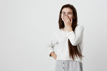 Happy dark-haired girl closing mouth with hand going to see surprise prepared by boyfriend standing and smiling in anticipation for something wonderful. Young cheerful lady covering face with hands