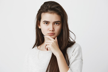 Fototapeta na wymiar Human emotions, feelings, reaction and attitude. Beautiful girl in casual t-shirt with long dark straight hair, keeping hand on chin in doubt and suspicion, feeling sceptical about something
