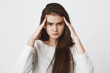 Photo of thoughtful serious concentrated brunette female model keeps fingers on temples, tries to remember important information, can`t find quick answer, poses against gray studio wall.