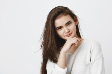 Young brunette female with oval face, dark appealing eyes having hair loose dressed in long-sleeved t-shirt holding hand under chin looking with dreamy expression at camera, thinking about something
