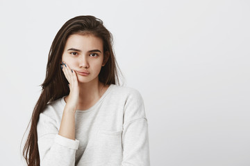 Obraz premium Beautiful young dark-eyed female with dark long hair wearing loose sweater, keeping hand on cheek, looking with serious expression at camera, isolated against gray background. Beauty and youth