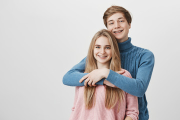 Love, happiness, and relationships concept. Happy caucasian male with fair hair dressed in blue sweater holding tight beautiful blonde Caucasian girlfriend. Young couple expressing positive emotions