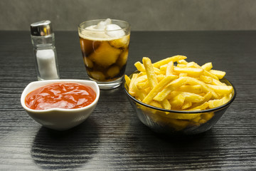 Salted french fries, ketchup and a glass of cola with ice on a wooden table.