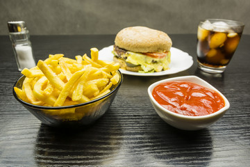 French fries and ketchup in bowls. In the background a beef burger and cola with ice in a glass.