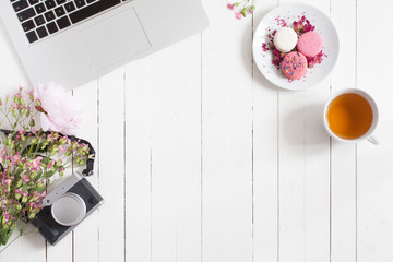 Feminine flat lay workspace with laptop, cup of tea, retro camera, macarons and flowers on white...