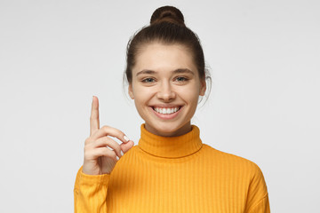 Attractive young woman in yellow sweater pointing up with her finger isolated on gray background