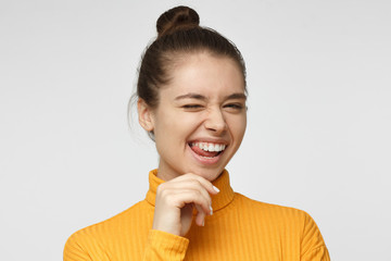 Beautiful girl in bright yellow polo neck sweater smiling and winking, looking at camera isolated on gray background