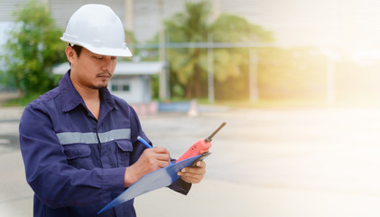 Fototapeta premium Asian engineer in safety uniform and white helmet taking note on clipboard on blurred industry plant background
