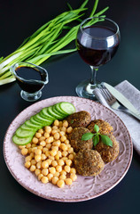 Tasty meat cutlets, chickpeas, fresh cucumber on a ceramic plate and a glass of wine on a black background.