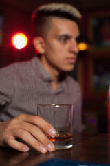 Blurry young hipster man sits at bar counter and holds glass of whiskey after long working day
