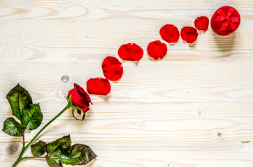 A path of petals leading from a red rose to a box with an engagement ring on a white wooden surface.
