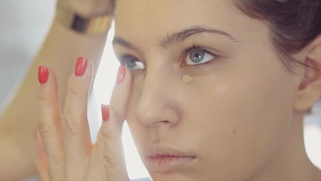 a portrait of a young and pretty woman whom a makeup artist paints, an artist applies a corrector under his eyes, a make-up artist edits the tone of his face