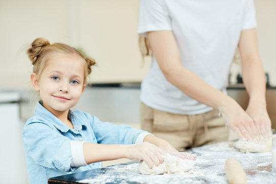 Pretty little girl kneading dough on table while halping her mother about the kitchen
