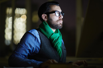 Serious man concentrating on browsing in the net while sitting at night in front of computer monitor