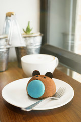 Brown bear cake with blue macaron nose with cup of coffee on wooden table at cafe. Blurred plants and window in background. Text space