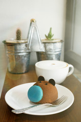 Brown bear cake with blue macaron nose with cup of coffee on wooden table at cafe. Blurred plants and window in background. Text space