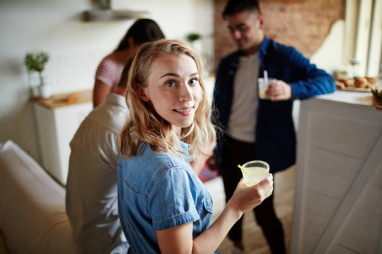 Pretty blonde with glass of drink looking at camer aamong her friends at home party