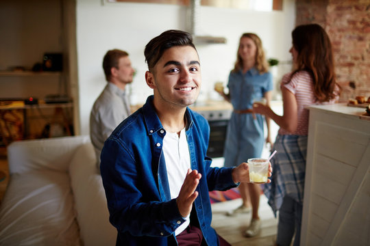 Smiling guy with glass of homemade drink and his friends on background enjoying home party