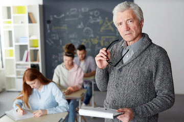Mature teacher with eyeglasses and notepad looking at camera with students writing test on background