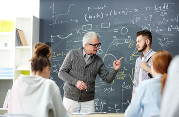 Strict grey-haired teacher pointing at one of students while talking to him by blackboard