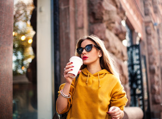 Stylish happy young woman wearing boyfrend jeans, white sneakers bright yellow sweatshirt.She holds coffee to go. portrait of smiling girl in sunglasses she greets friends