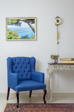 Vintage Furniture - Interior composition of retro blue armchair, vintage wooden beige table, and pendulum clock over off white wall, tiled beige floor and orange ornate carpet