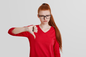 Portrait of pretty young female shows dissaproval and dissagreement, keeps thumb down, has opposite...