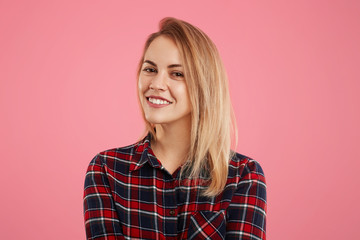 Fototapeta premium Pretty adorable woman with pleasant smile looks directly into camera, dressed in checkered shirt, isolated over pink background. Smiling friendly young female rejoices her success, poses indoor