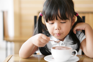 Asian children cute or kid girl holding spoon and blow for delicious hot cocoa or chocolate drinking in white cup and sloppy for breakfast in the morning on table at home or cafe restaurant