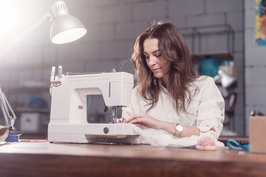 Attractive Caucasian seamstress working stitching with sewing machine at her workplace in studio loft interior