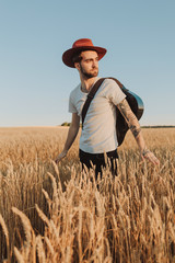 Young musician in hat with guitar walking on a summer wheat field. Bearded stylish young man.