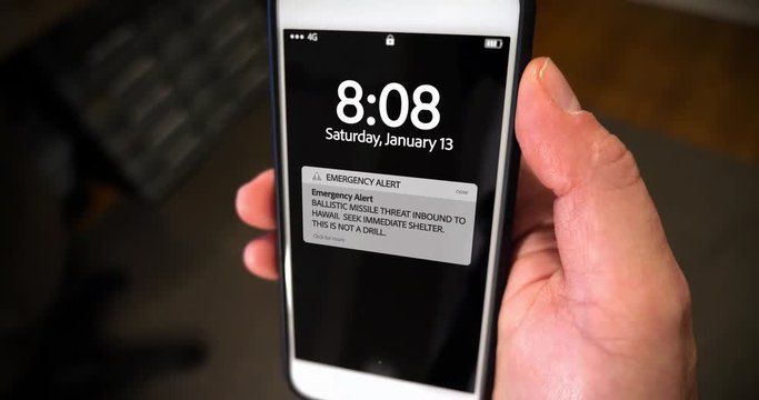 A man holds a smartphone showing an incoming ballistic missile alert notification. Screen contents simulated on a generic device.	