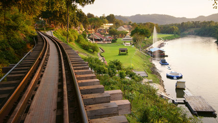 Naklejka na ściany i meble nature landscape railroad tracks and fountain on river with green grass meadow in the resort or hotel for tourist travel on relax holiday with warm sunlight at kanchanaburi in Thailand