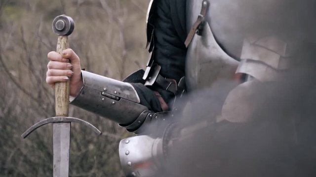 A woman in armor leans on a sword sitting by the fire