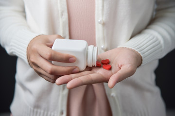 Female hands is holding a white pill bottle and red pills in heart shape.
