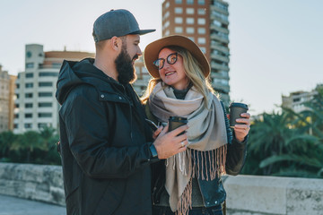 A couple of young tourists, friends are walking along the city street, drinking coffee while they are happily looking at each other. Vacation, travel, adventure, lifestyle. Blurred background.