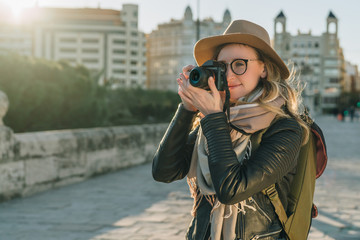 Sunny day, autumn. Young woman tourist, photographer, hipster girl dressed in hat and eyeglasses, stands on city street and takes photo. Vacation, travel, adventure, sightseeing. Blurred background.