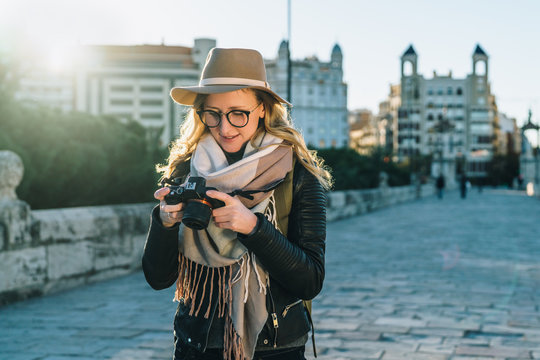 Backlight. Young smiling woman tourist, photographer, hipster girl dressed in hat and eyeglasses, stands on city street and uses camera, looks images on screen. Vacation, travel. Blurred background.