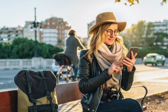 Sunset. Young woman tourist with backpack and camera, dressed in hat and glasses, sits on bench in city street and uses smartphone. Hipster girl chatting, blogging. Vacation, travel, social networks.
