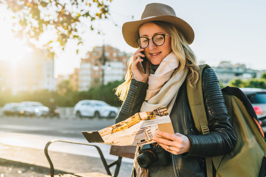 Young woman tourist with backpack,dressed in hat and glasses,sits on bench in city street, looks at map and talks on her cell phone.In background,in soft focus, road and car.Hipster girl using gadget.