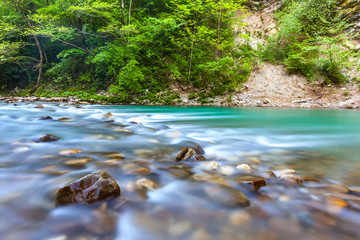 Wet brown stones in a transparent mountain smooth river of turquoise color against a rock background with green trees. Yew and Boxwood Grove, Adler, Sochi, Russia.