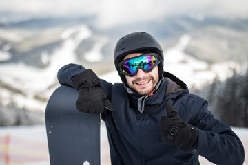 Man snowboarder standing on the top of the snowy slope with snowboard, smiling to the camera,...