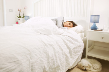 Woman lying in bed next to dog happy looking camera