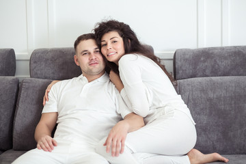 Obraz premium Portrait of young couple sitting together on grey sofa in modern white apartment or domestic room. Happy lovely family embracing and hugging together close-up