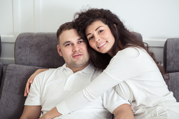Portrait of young couple sitting together on grey sofa in modern white apartment or domestic room. Happy lovely family embracing and hugging together close-up