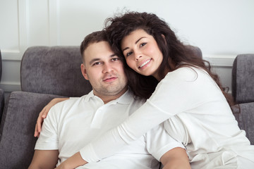 Obraz premium Portrait of young couple sitting together on grey sofa in modern white apartment or domestic room. Happy lovely family embracing and hugging together close-up
