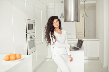 Fototapeta na wymiar Portrait of a young pretty woman with long black curly hair at home kitchen with laptop on background at morning