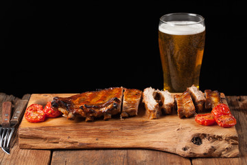 Pork ribs in barbecue sauce and honey baked tomatoes on the old wooden table. Meats and light beer on black background with copy space