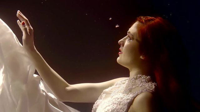 A young woman with red hair touches the fabric of her dress and pulls her arm forward under the water