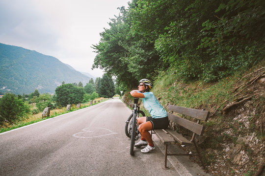young girl in helmet and sports clothes sitting resting dreams and looking out into the distance on a wooden bench on a cycling route in Italy in mountainous area.On the asphalt the heart is chalked